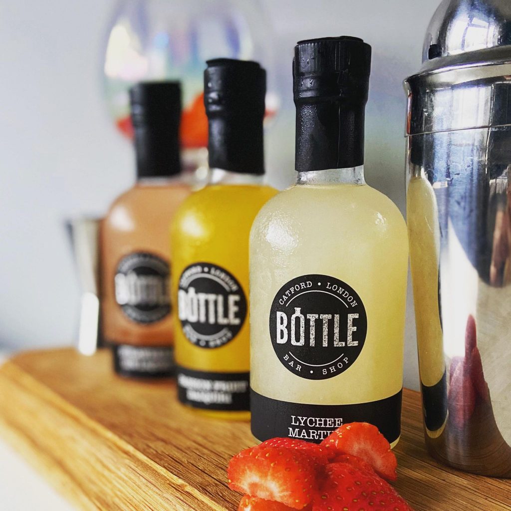 A picture of 3 cocktail bottles from bottle bar and shop with a cocktail mixer in the foreground and an empty gin glass in the background.